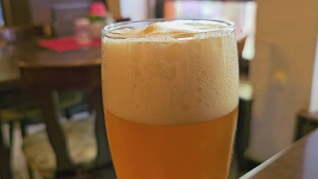 Fresh draft beer in a glass with creamy foam head, golden amber liquid, condensation droplets, soft bar lighting, and shallow focus restaurant background for brewery or nightlife video.