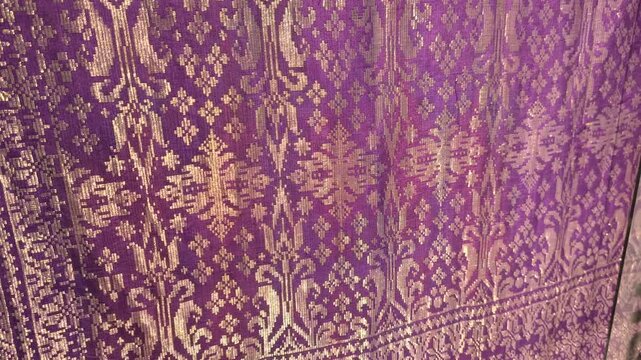 Detailed close-up of luxurious Songket fabric. The intricate gold embroidery shines across the handwoven surface. This prestigious Malaysian textile is reserved for the most special occasions.