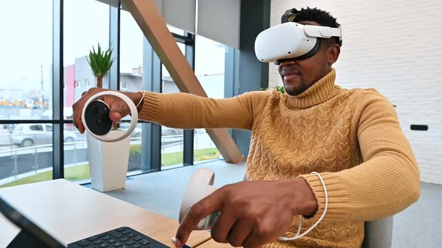 Young african american man wearing virtual reality headset using controllers to explore digital simulation while sitting at office desk.