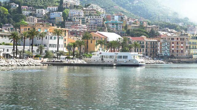 Aerial footage of Rapallo, Italy, showing sparkling harbor water, palm-lined promenade, and vibrant hillside houses under soft daylight. Wide scenic view with no people visible.