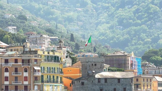 Bright, wide angle footage of historic Rapallo Castle with the Italian flag waving, colorful Ligurian buildings, and lush green hills in soft daylight. No people are visible. (196 characters)