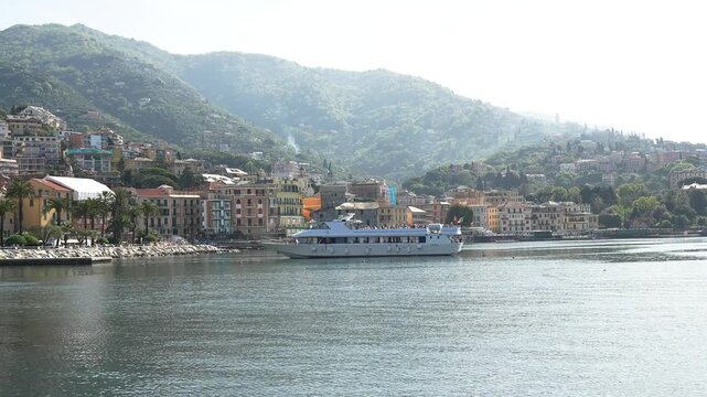 Wide shot of Rapallo, Italy, featuring a ferry gliding on calm water by vibrant buildings and green hills under bright daylight. Beautiful Italian Riviera coastal scenery, steady camera, crisp illumin