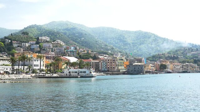 Wide aerial shot of Rapallo, Italy, showcasing colorful waterfront buildings, lush green hills, palm trees, and tranquil bay under bright, clear daylight. Vibrant, scenic, and inviting drone video.