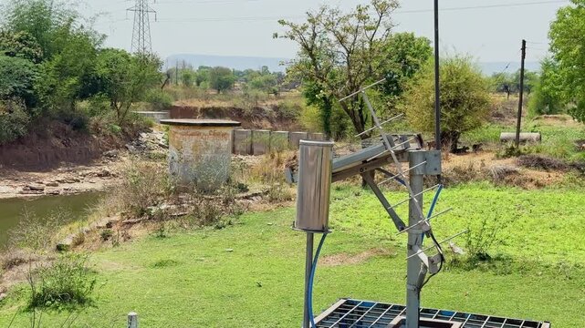Closeup of solar powered monitoring equipment and antenna installation near a rural riverbank surrounded by green vegetation and dry landscape!
