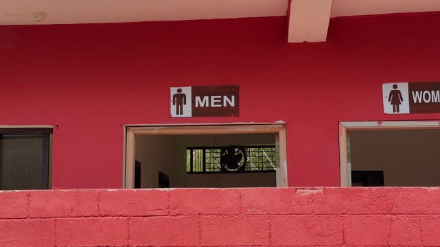Panning shot showing men and women restroom entrance signs on a bright red exterior wall of a public building in daylight with simple architectural framing!