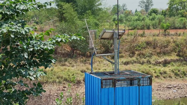 Small blue solar powered monitoring station with antenna and solar panel installed near a rural water body surrounded by dry vegetation!