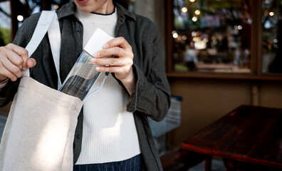 Woman placing reusable water bottle into canvas tote bag outdoors. Sustainable lifestyle, zero...