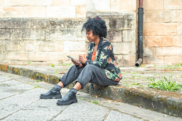 Fototapeta premium African American adult woman sitting on the street steps calmly drinking coffee and using her mobile phone