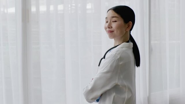 Asian Female Doctor in White Coat Smiling with Arms Crossed by Bright Window