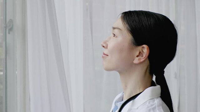 Asian Female Doctor in White Coat Looking Out of Windo