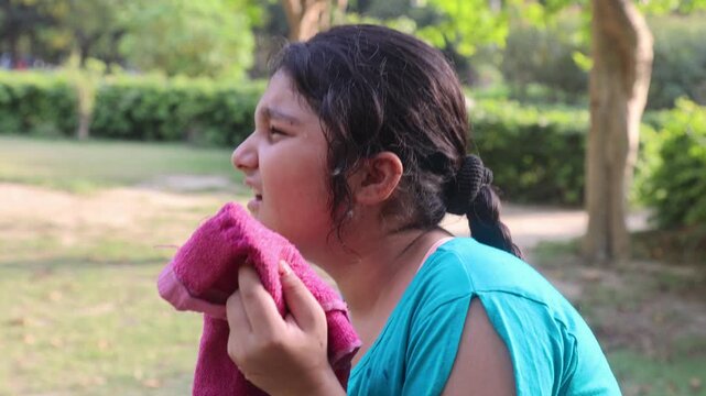 Young Indian girl wiping sweat with a towel during intense summer heat in an outdoor park, expressing discomfort, tiredness, and the effects of high temperature weather in natural surroundings.