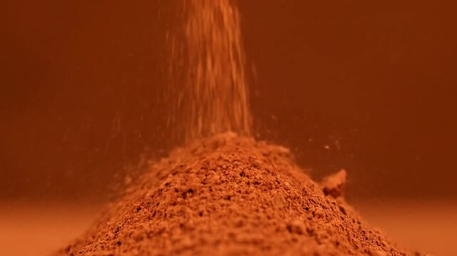 Fine reddish-brown powder falling onto a growing pile against a solid warm background. Macro shot showing the texture of the spice or pigment particles as they settle into a heap.