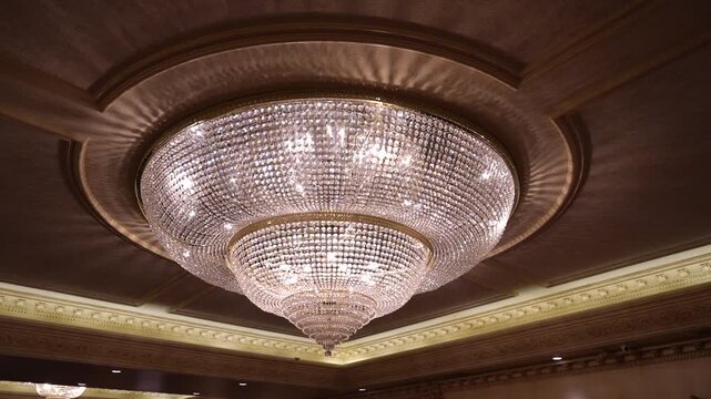 Large crystal chandelier glows brightly with intricate light refraction patterns suspended from a detailed ornate ceiling in this indoor photograph.