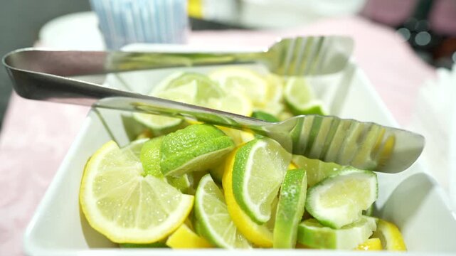 Fresh yellow lemon and green lime wedges sit inside a white dish accompanied by metal tongs in this bright food photograph.