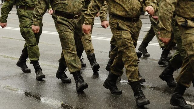 Camouflage-clad soldiers march in close formation on a rainy city street, then the column passes, revealing an empty wet roadway with spectators standing behind metal barriers