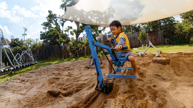 Cute Asian boy playing with sand excavator toy in outdoor playground sandbox. Active childhood, learning and recreational activity in sunny outdoor environment.