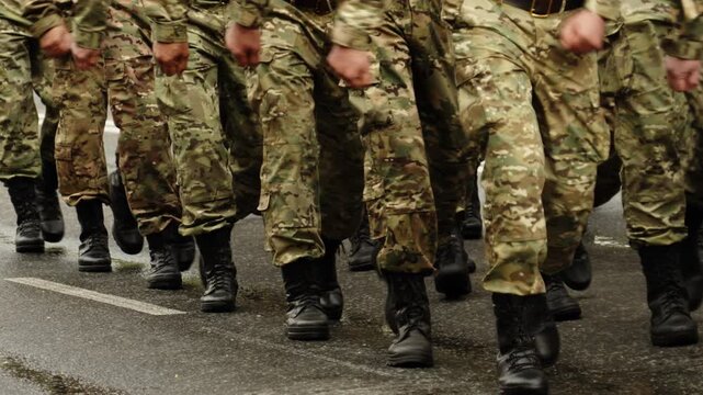 Close-up view of uniformed soldiers marching in synchronized formation on a wet asphalt road during a military parade, showing camouflage fatigues, black combat boots, and cadence.