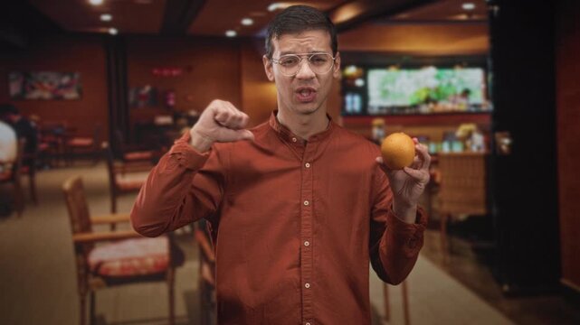 Young hispanic man holding orange and showing thumb down gesture in building cafe interior; taste test disapproval.
