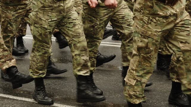 Close-up of uniformed troops in camouflage combat boots marching in formation along a rain-soaked street, followed by an empty roadway with reflective puddles after the march