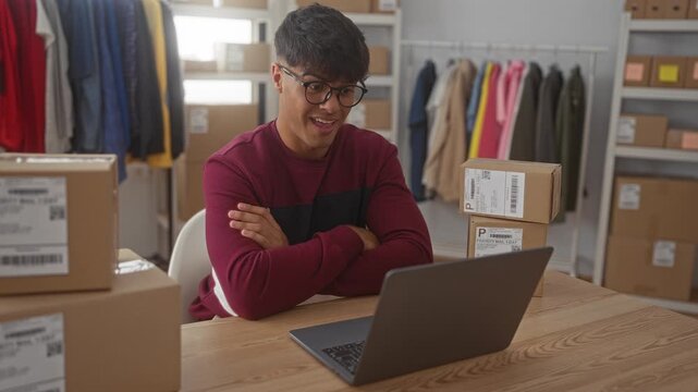 Man smiles and fist pumps at laptop amid stacked shipping boxes and clothing racks in studio workspace; business joy.