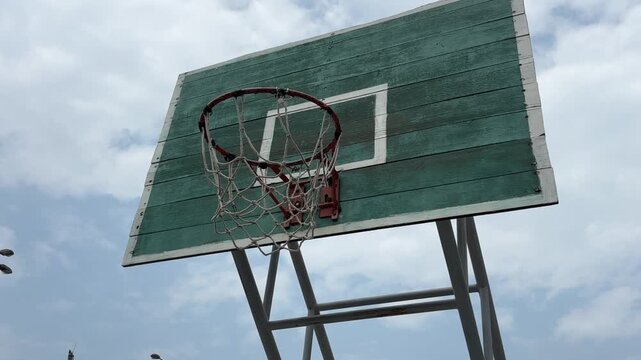Old outdoor basketball hoop on a green wooden backboard under a clear blue sky. Tilt up shot