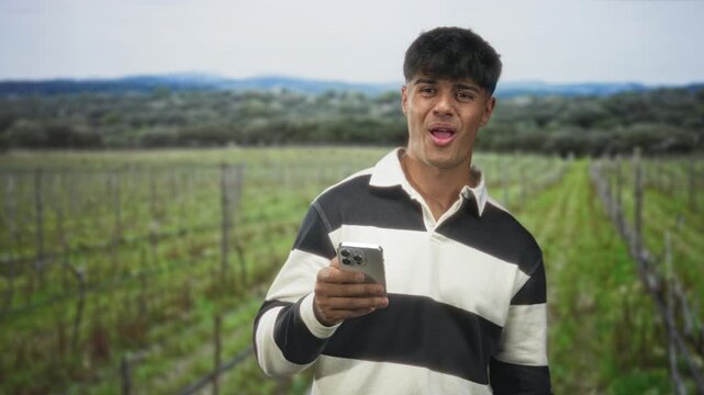 Young hispanic man holding smartphone and glancing at the screen while making a fist pump gesture in a vineyard; achievement joy.