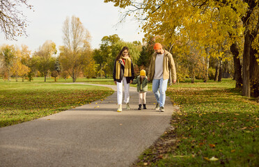 Fototapeta premium Family walking together in autumn park path. Parents and child stroll a route under golden leaves, holding hands and enjoying fresh air on a sunny day. Concept: family unity and shared joy.