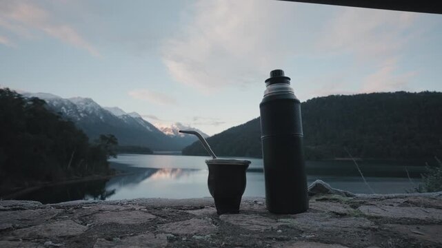 A mate cup and thermos sit on a stone ledge overlooking a calm lake in Patagonia. Ideal for travel content or cultural documentaries.