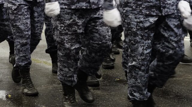 Close-up view of uniformed personnel in blue camouflage marching in formation with black combat boots on a wet street, finishing with a single step as the column passes.