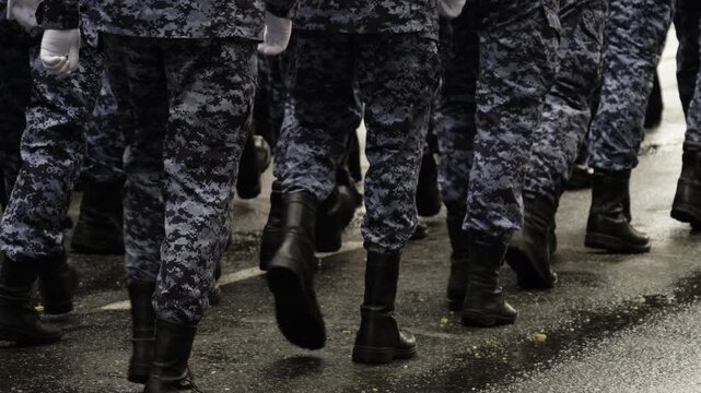 Close-up view of uniformed soldiers in camouflage marching in tight formation along a wet urban street, black boots lifting in unison during a disciplined parade drill under rain