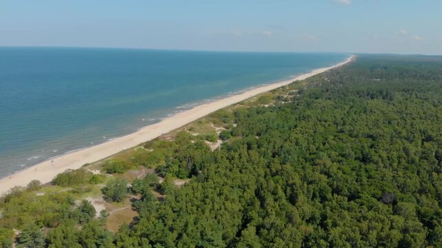 Drone shot capturing a long, pristine sandy beach and the calm blue waters of the Baltic Sea, bordered by a dense green pine forest. The footage showcases the natural beauty and coastal landscape of t