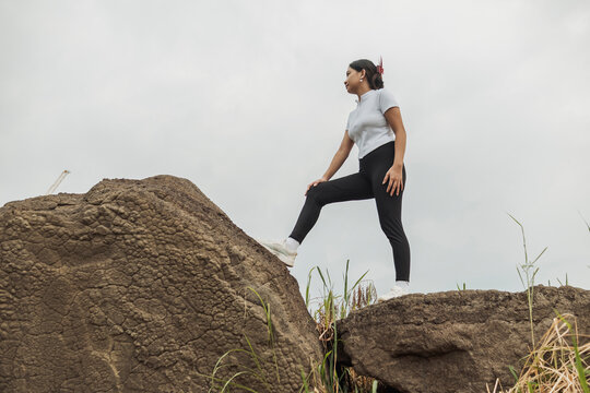 Young Southeast Asian woman standing on top of a large rock while admiring the outdoor scenery during a hiking adventure.