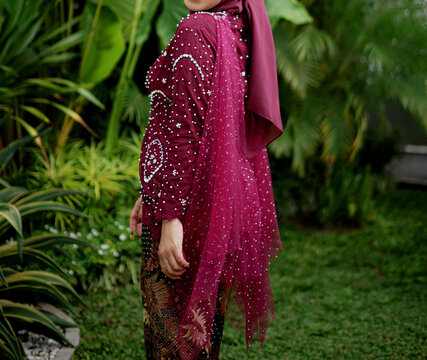 A faceless female body wearing a luxurious burgundy kebaya with beautiful batik cloth, standing in a green garden.