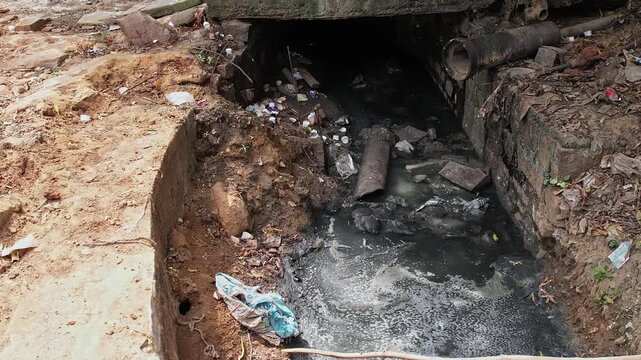 Wide view shows a heavily polluted open drain filled with debris sewage and plastic waste