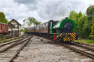 Fototapeta premium Vintage steam locomotive, 2623 Hawarden built in 1940, with maroon carriages on a heritage railway track by a traditional wooden signal box under a smoky, overcast sky. 