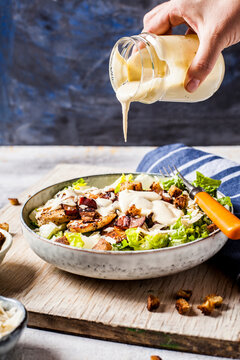 Hand of person pouring dressing over bowl of Caesar salad with romaine lettuce, Parmesan cheese, bacon, chicken breast and croutons