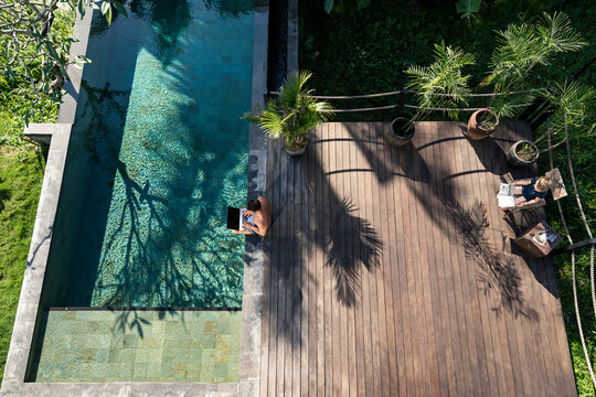 Mature man sitting at the poolside, using laptop, while daughter is reading