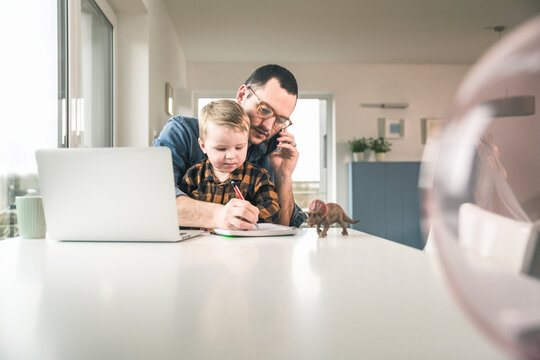 Busy father working at table in home office with son sitting on his lap