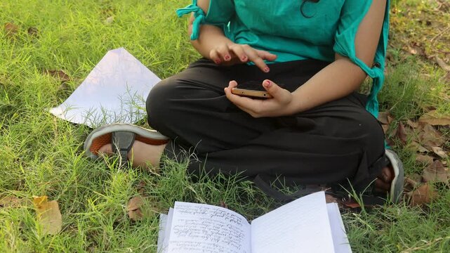 Close view of a young Indian girl scrolling on smartphone and searching study related information while sitting on grass with open books during hot summer weather in a green park in India.