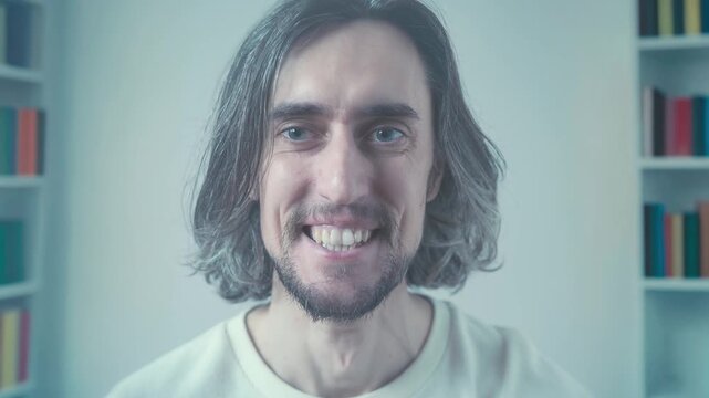 A man with medium-length brown hair and beard calmly sits before colorful blurred books background, transitioning from neutral to broad smile joyful eyes thoughtful expression in soft pastel lighting