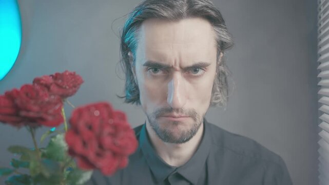 Comical guy trying to be serious, a man sitting indoors against a muted grayish-blue background vibrant red roses, showing changing facial expressions from calm to contemplative under soft lighting