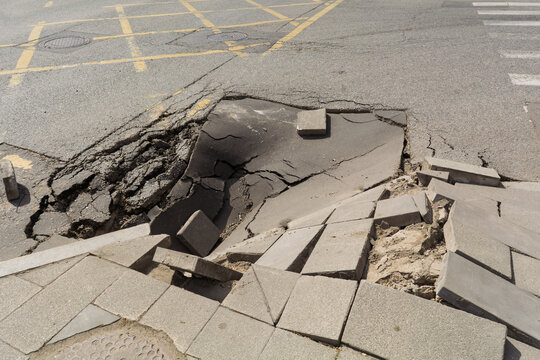 Collapsed asphalt and broken sidewalk blocks expose dangerous road damage at urban intersection after pavement failure near crosswalk