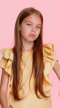 Young child girl frowns shows a thumbs down gesture clearly expressing disappointment, rejection, frustration. School kid isolated on pink background portraying strong negative emotion with confidence