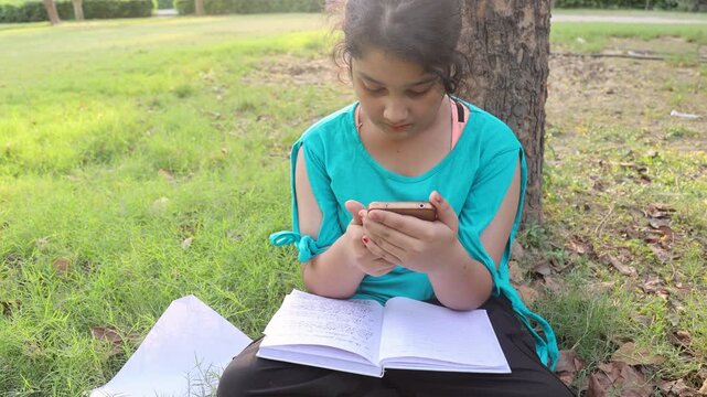  Young Indian girl sitting under a tree in a green park while using a smartphone and studying from an open book during hot summer weather. Peaceful outdoor learning and lifestyle moment in