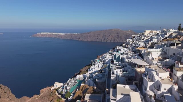 Drone footage revealing picturesque houses on the edge of Santorini caldera with Oia in the background, showcasing iconic cliffside architecture and the dramatic Aegean island landscape.