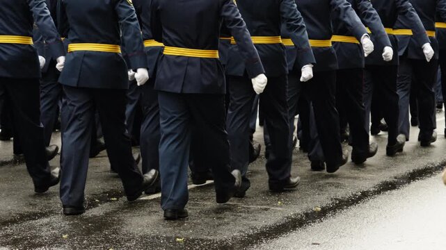 Uniformed marching unit in blue dress attire with yellow belts advances in tight formation along a wet street, synced strides and polished boots show disciplined parade cadence
