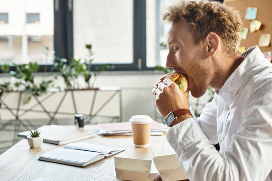 Young man enjoying a delicious burger while working in a cozy modern workspace