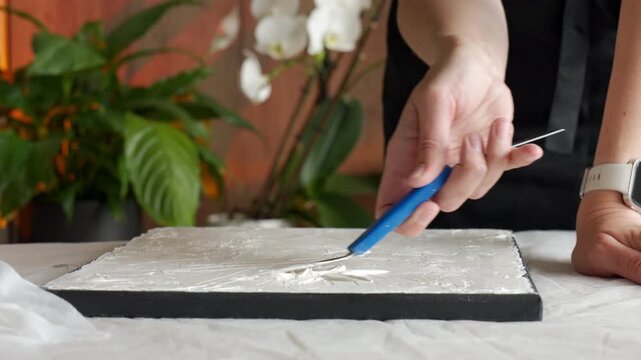 Artist creates textured artwork on canvas at home, spreading white plaster paste with a palette knife at a covered work table. Concept of DIY wall decor and creative hobby.