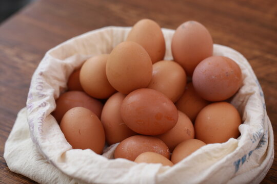 A high-angle, close-up shot shows a cluster of smooth, brown chicken eggs packed inside a cream-colored textile pouch. The bag is gathered at the edges, resting on a warm, dark wood surface. The light