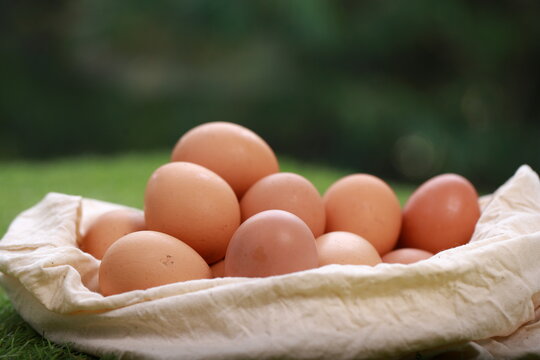 A high-angle, close-up shot shows a cluster of smooth, brown chicken eggs packed inside a cream-colored textile pouch. The bag is gathered at the edges, resting on a warm, dark wood surface. The light